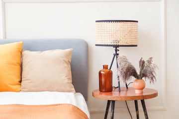 Stylish lamp on wooden nightstand next to flower in big glass vase in scandinavian bedroom interior