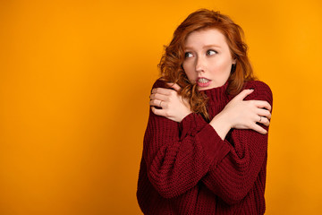 Beautiful red-haired girl in a burgundy sweater looks away clasping her shoulders with her hands.