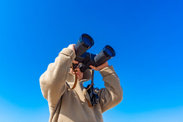 A boy with binoculars on a background of blue sky looks up.