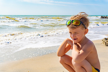 A joyful and happy boy in sunglasses on his head sits on the sea sand on the beach, smiles and looks around.