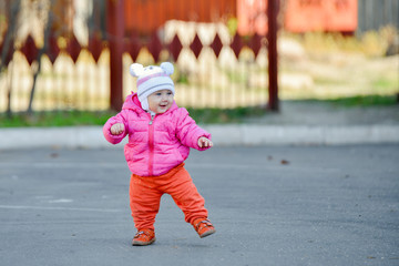 Fototapeta premium A little girl in a red jacket learns to walk on the road in the fall afternoon.