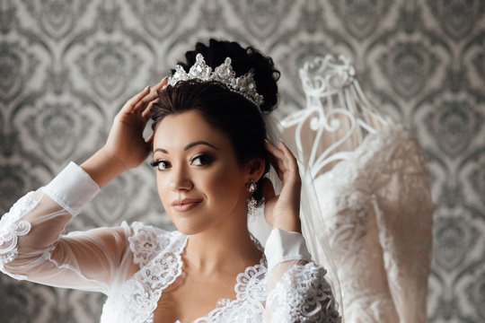 Beautiful happy brunette bride in a stylish robe posing near manequin wedding dress, morning wedding preparation concept, bride portrait face closeup sensual make up and diadem on her head.