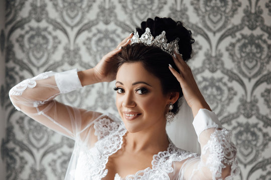 Beautiful happy brunette bride in a stylish robe posing near manequin wedding dress, morning wedding preparation concept, bride portrait face closeup sensual make up and diadem on her head.