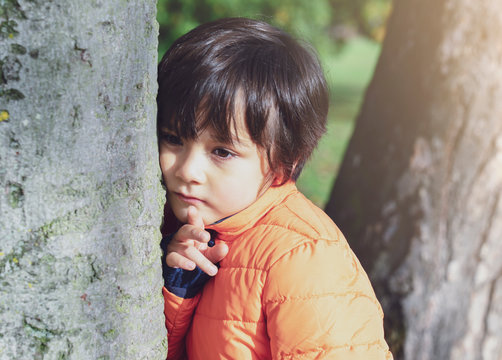 Portrait of sad kid standing next to big tree, Lonely child standing alone and looking down with thinking face in the park,Little boy with worrying face looking down, lost children or homeless kid