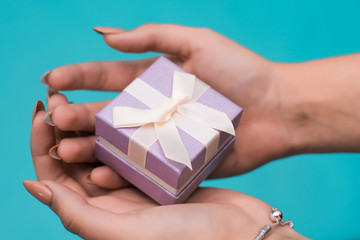 Close up shot of female hands holding a small gift wrapped with pink ribbon. Small gift in the hands of a woman indoor on blue background. focus on the little box.