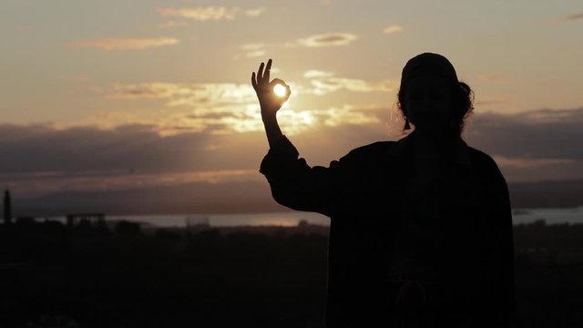 Silhouette of a hipster girl standing in the sunset with ohm sign with her fingers, meditating  