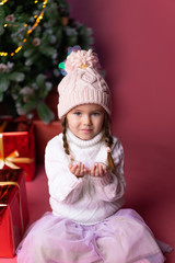 Beautiful little girl in the hat playing with snow near presents and christmas tree. Bokeh lights on the background. Concept christmas