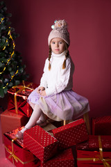 Beautiful little girl in the hat sitting near presents and christmas tree. Bokeh lights on the background. Concept christmas