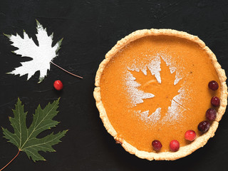 Homemade traditional American pumpkin pie on a black background. Decorated with powdered sugar in the form of a leaf and cranberries. Near autumn maple leaves. Top view.