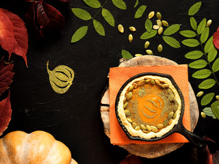 Homemade traditional American pumpkin pie located in a cast-iron shape. Decorated with pumpkin seeds. On a black background colorful autumn leaves and pumpkin. Top view.