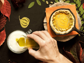 Homemade traditional American pumpkin pie located in a cast-iron shape. Decorated with pumpkin seeds. On a black background colorful autumn leaves. Girl's hand pours a cup of hot matcha tea. Top view.
