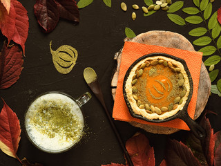 Homemade traditional American pumpkin pie located in a cast-iron shape. Decorated with pumpkin seeds. On a black background colorful autumn leaves and a cup of hot matcha tea. Top view.