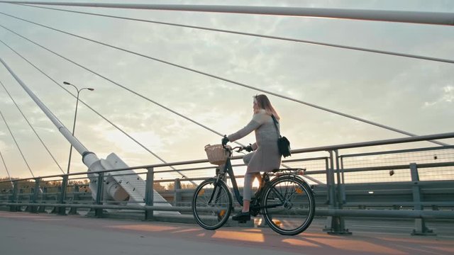 Young Woman is Riding Bicycle or Cycling Bike on Suspension Bridge in Autumn Sunny Morning. Urban Cycle Chic and Ecological Transportation. Amazing 4K Long Tracking Background Shot with Copy Space