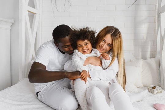 Portrait of happy multiracial young family lying on cozy white bed at home, smiling international mom and dad relaxing with little biracial girl child posing for picture in bedroom
