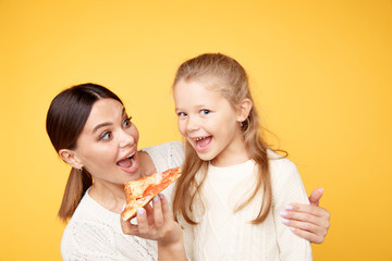 Mother and daughter eating pizza together and having fun isolated over the yellow studio