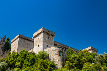 Fototapeta na wymiar The medieval castle of the ancient village of Narni. Umbria, Terni, Italy. The blue sky in summer. The stone walls and towers of the fortress. Green trees, vegetation, nature.