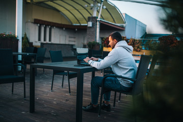 Young freelancer in gray hoodie uses laptop sits in cafe and reads news, checks e-mail. E-news concept.