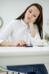 Young brunette in a white shirt is waiting for a business partner in a cafe. Woman at a business meeting. Girl drinks coffee during the break and uses a smartphone reads sms.