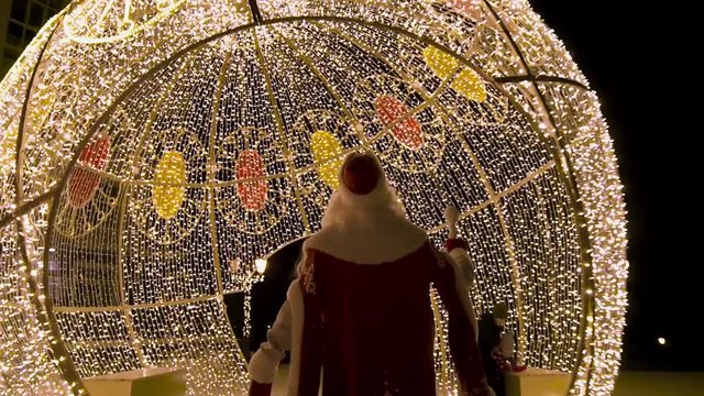 Rear view of Santa Clous coming to the Christmas lights decorations in the street at night, winter holidays concept. Clip. Amazing spherical construstion covered with garlands.