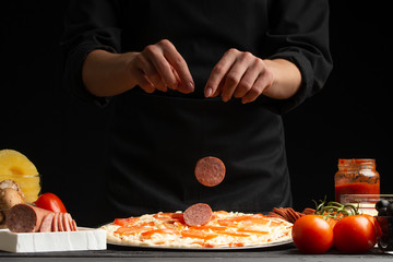 The chef cooks Italian pizza, sprinkled with slices of salami. Freeze in the air. Against the background of pizza ingredients. Black background, horizontal frame.