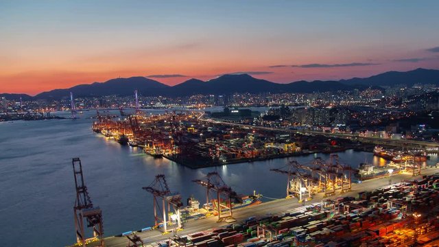Timelapse illuminated Busan harbour with large operating cranes at waving water reflecting evening sun light panning up