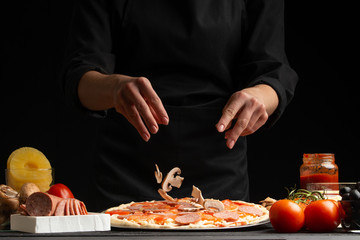 Chef cooks Italian pizza, sprinkles with mushrooms. Freezing in motion. Against the background of pizza ingredients. Black background.