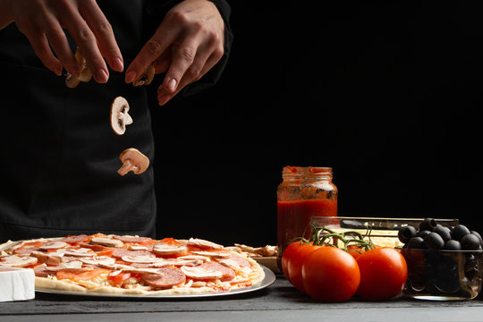 Chef cooks Italian pizza, sprinkles with mushrooms. Freeze in motion. Against the background of pizza ingredients. Black background with space for design.
