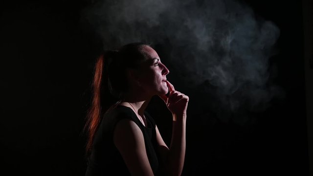 A pensive brunette is in a cloud of smoke. Portrait of a woman smoking a vape behind her, soaring. Black background. Passive smoking. Bad habits.