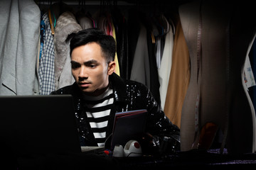 Black Sequin jacket of Asian Fashion Designer Man checks order and sale growth on Notebook computer. Gay tailor designs new trend of collection to Cloth rack backgrounds, low key exposure copy space