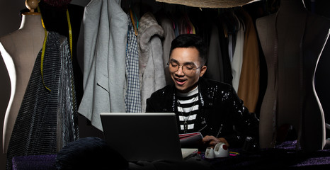 Black Sequin jacket of Asian Fashion Designer Man checks order and sale growth on Notebook computer. Gay tailor designs new trend of collection to Cloth rack backgrounds, low key exposure copy space