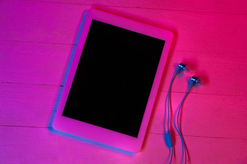Top view of set of gadgets in purple neon light and pink background. Tablet with black screen and headphones. Copyspace for your advertising. Tech, modern, gadgets.