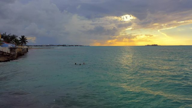Aerial: Golden Sunset Behind Clouds Over Tropical Water, Three People Swimming Near Waterfront Homes - Nassau, Bahamas