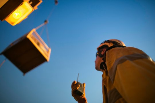 Defocused picture of rigger crane operator miner wearing helmet, using two way safety radio communicating with crane operator while load is being lift at construction mine site Perth, Australia 