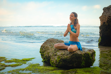 Young woman sitting on the rock, meditating, practicing yoga and pranayama with namaste mudra at the beach, Bali