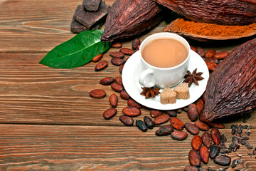 Cup of cacao drink with green leaf, cocoa pods, cacao powder, chocolate, sugar, cacao beans and star anise on a wooden background.