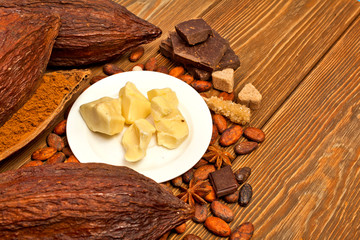 Cocoa butter in the plate, cacao powder, cocoa pods, chocolate, sugar, cacao beans and star anise on a wooden background.