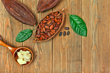 Cocoa beans in cacao pod with natural green leaves and cocoa butter in spoon on old wooden background. Top view. Text space.
