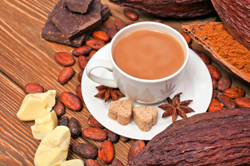 Cup of cacao drink with cocoa pods, cacao powder, cocoa butter, chocolate, sugar, cacao beans and star anise on wooden background.