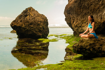 Young woman sitting on the rock, meditating, practicing yoga and pranayama at the beach, Bali....