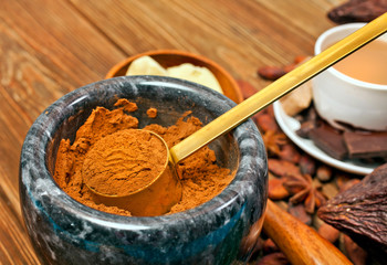 Cocoa powder in stone mortar with cup of cacao drink, cocoa pod, chocolate bar, cocoa beans and star anise on wooden background.