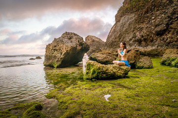 Woman meditating, practicing yoga and pranayama. Namaste mudra. Lotus pose. Rocks covered by green...