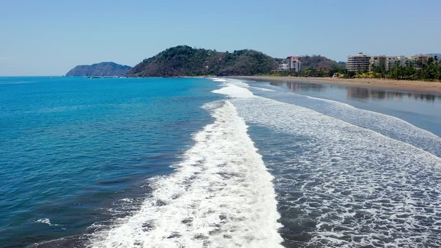 Aerial: Several People Wading Through Gentle Foamy Waves in Vibrant Blue Ocean With Lush Green Hills Behind - Jaco, Costa Rica