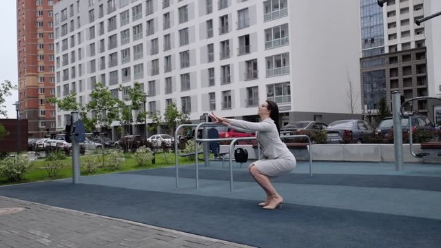 A woman in a business suit and heels does a sit-up outside. Business woman in glasses doing exercises during the break. The girl is engaged in fitness in the courtyard of the house on the playground.
