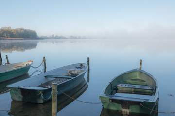 Fototapeta na wymiar on a lake some rowboats are moored and on the lake there is fog and the lake is quite calm