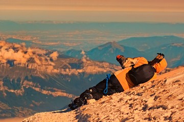 A man sits in a sleeping bag and drinking tea from a thermos on the background of winter mountains.