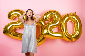 happy woman in holiday dress with new year 2020 balloons isolated over pink