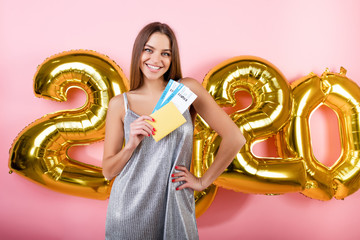 beautiful woman with plane tickets and passport with 2020 new year balloons isolated over pink