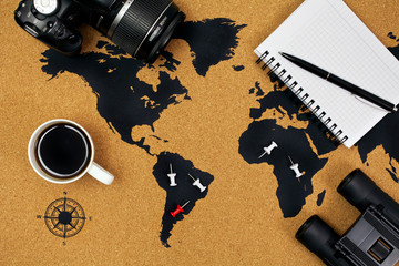Cup of black coffee on a map with pins, camera, binoculars, notepad and pen. Top view.