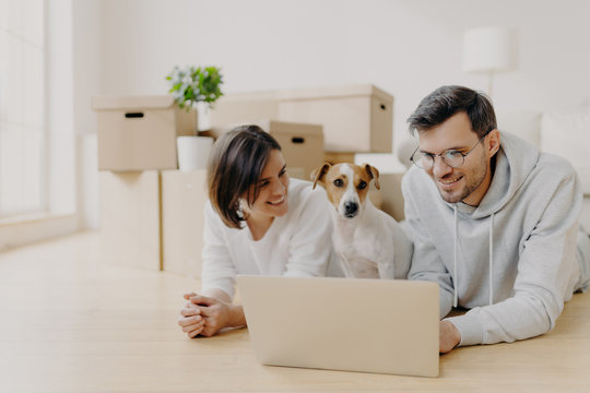 People and new apartment concept. Happy young husband and wife relax on floor, use modern laptop computer for surfing net, pose in their new home or flat, their dog poses near, have little rest