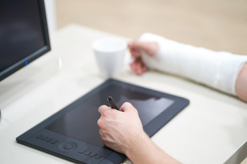 Fototapeta premium Artist with broken arm drawing something with graphic tablet. Hold pen in broken fractured arm in plaster cast.